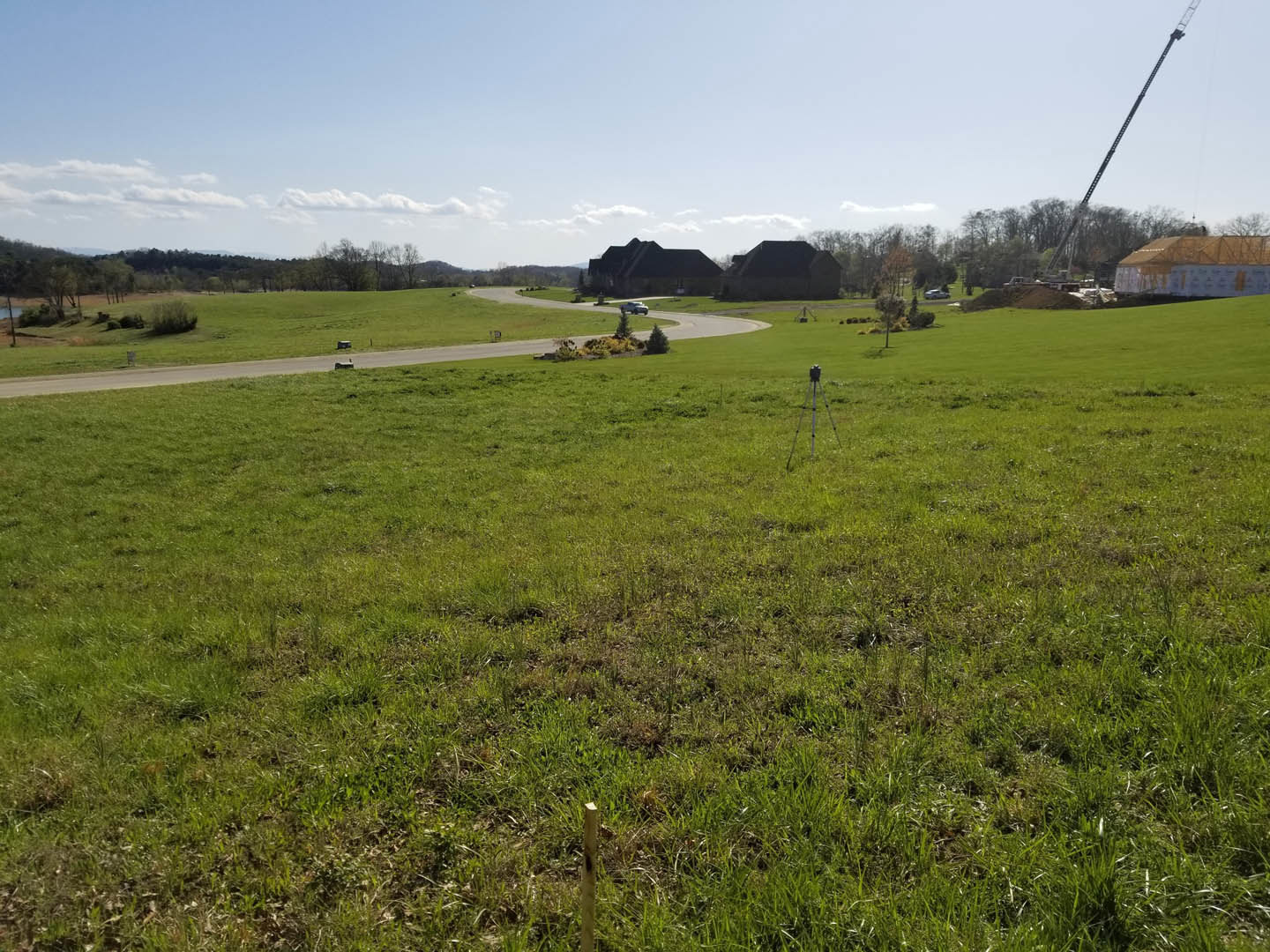 Wide grassy field bordered by a paved road, distant buildings with pitched roofs, scattered trees, and a clear blue sky with clouds overhead.