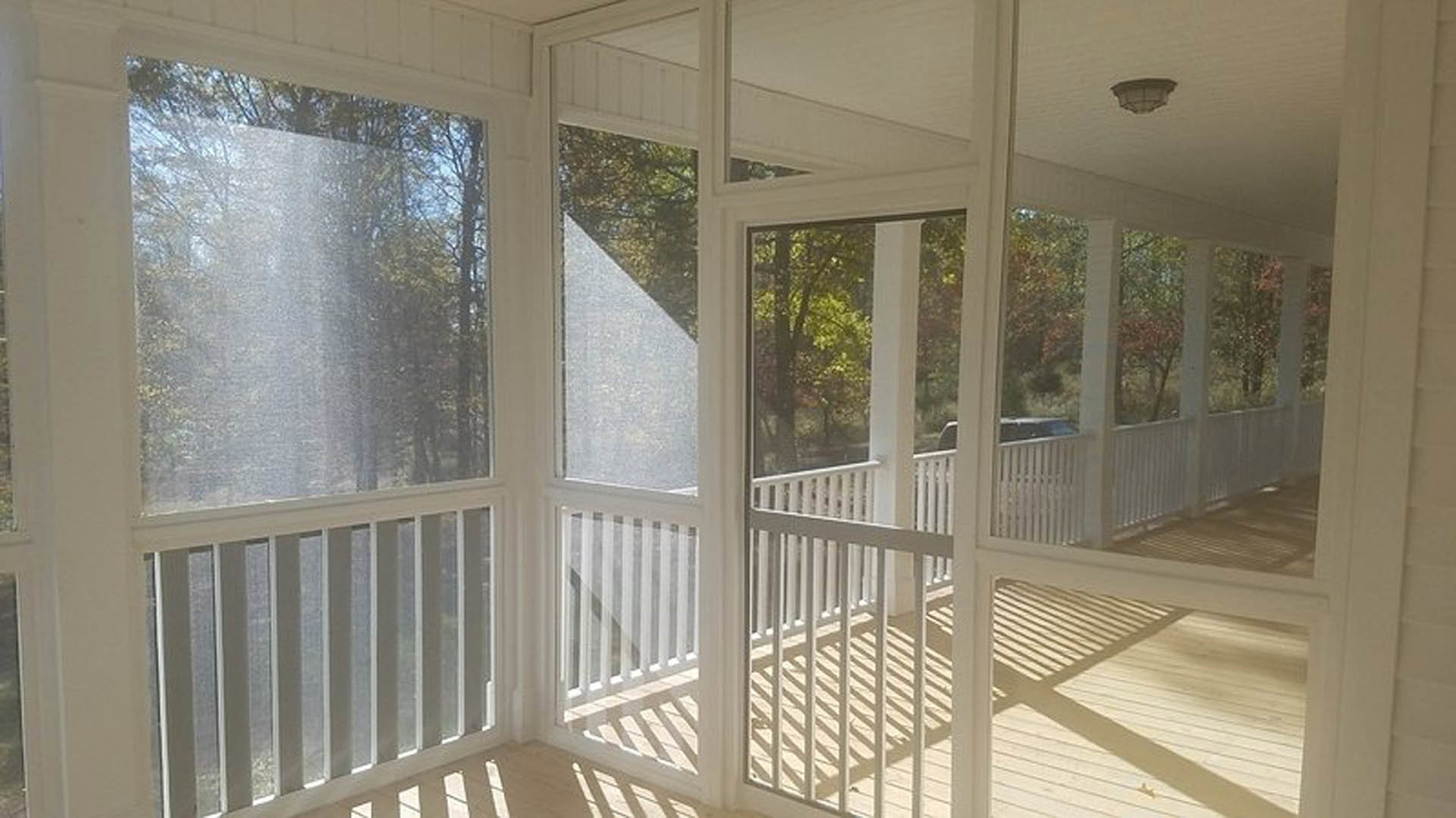 Wooden porch with screened door, white railing and balusters, wall-mounted light fixture, white-framed window overlooking trees, close-up of deck flooring