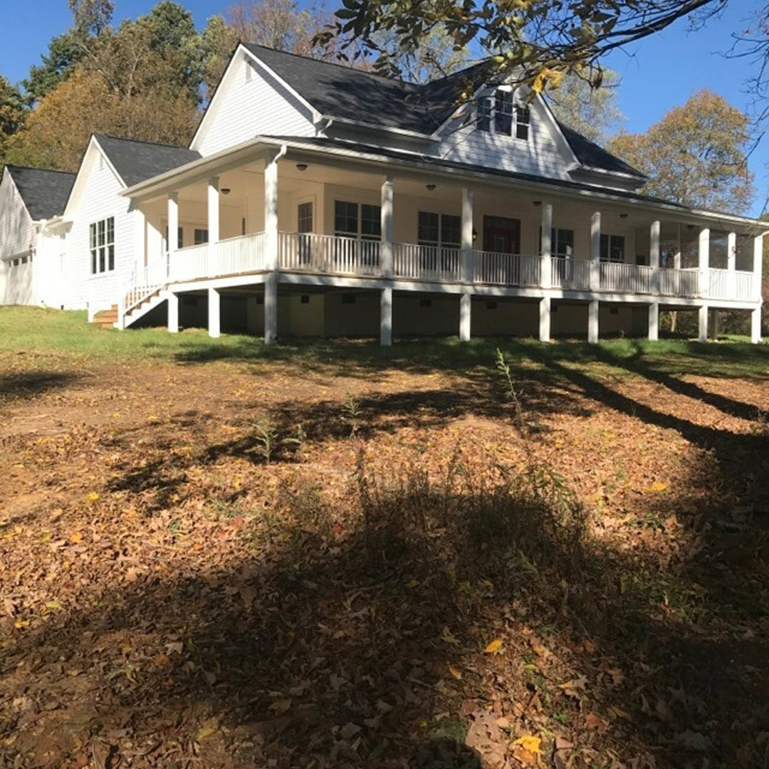 White two-story house with wide covered porch, white railings, multiple windows, grassy lawn scattered with leaves, tree branches framing the roofline