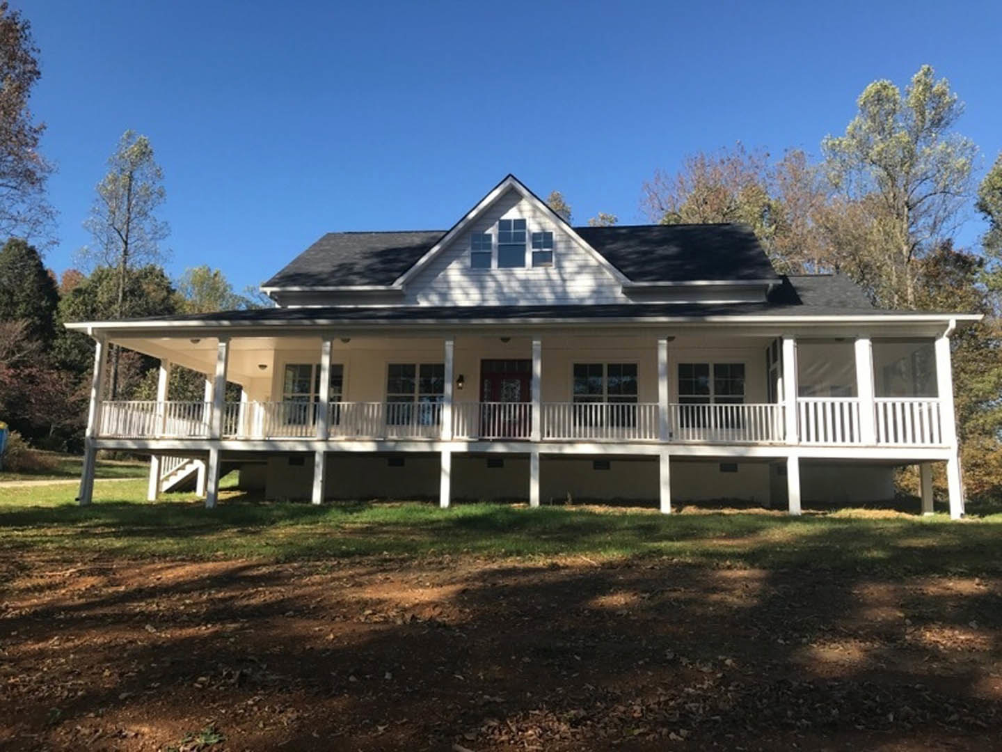 White siding home with covered front porch, white railings and posts, dirt walkway bordered by grass, multiple windows with white frames, gabled roof, trees and blue sky in