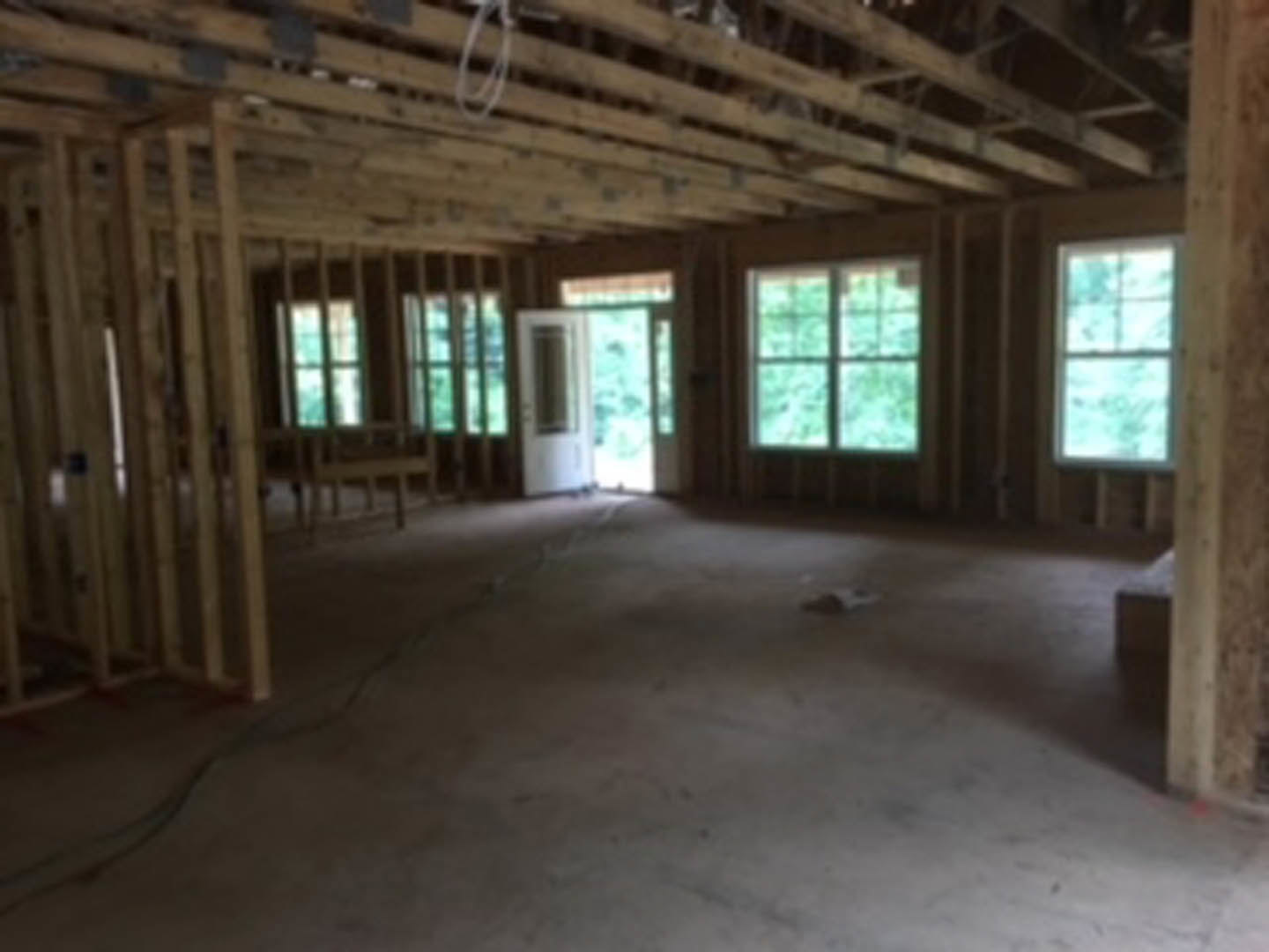 Sunlit room with wood plank flooring, exposed ceiling beams, multi-pane windows, and a wooden door, surrounded by natural daylight and green outdoor scenery.