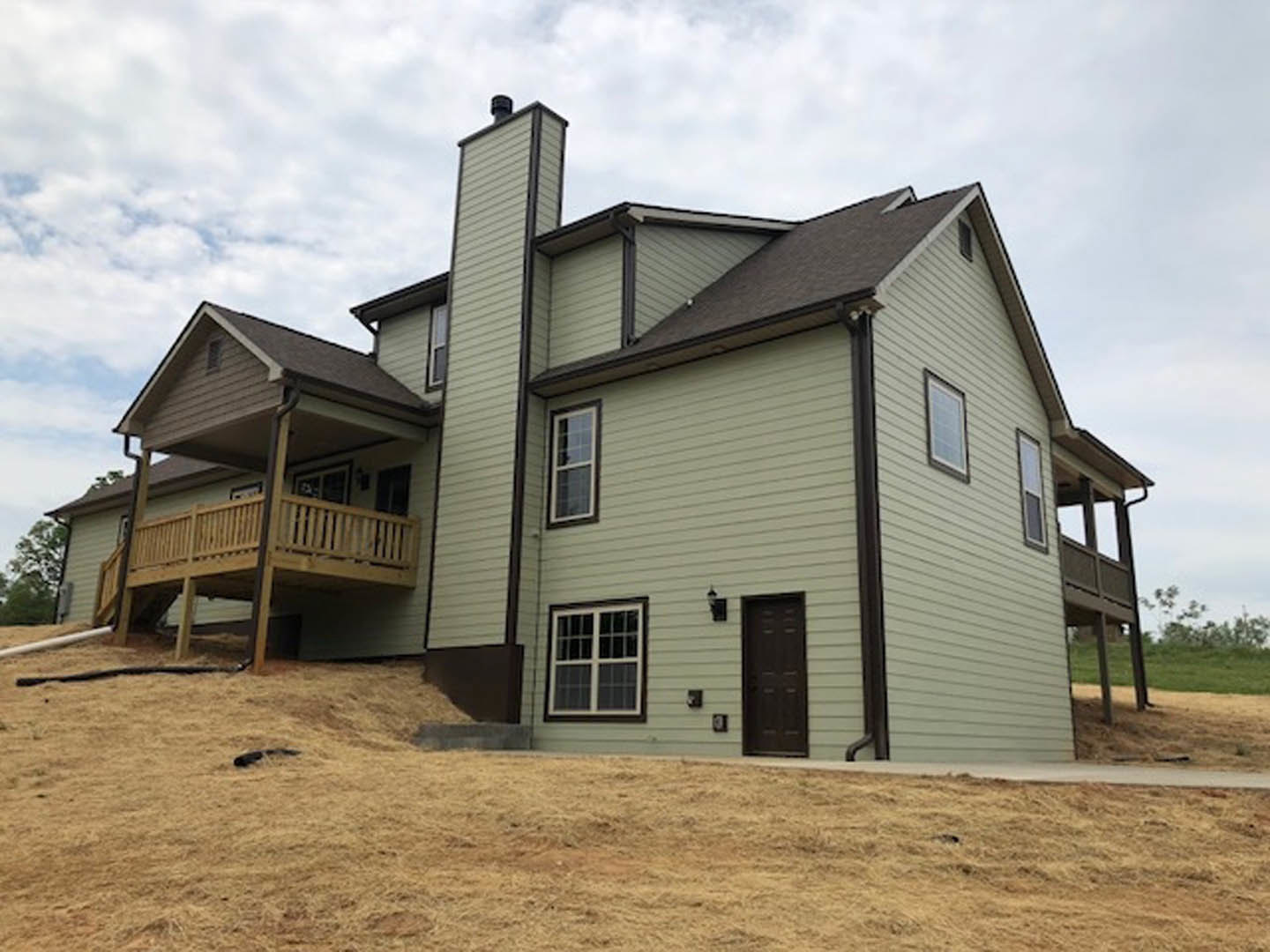 Two-story house with white siding, multi-pane windows, black front door with white trim, wooden side deck, and green lawn under partly cloudy sky