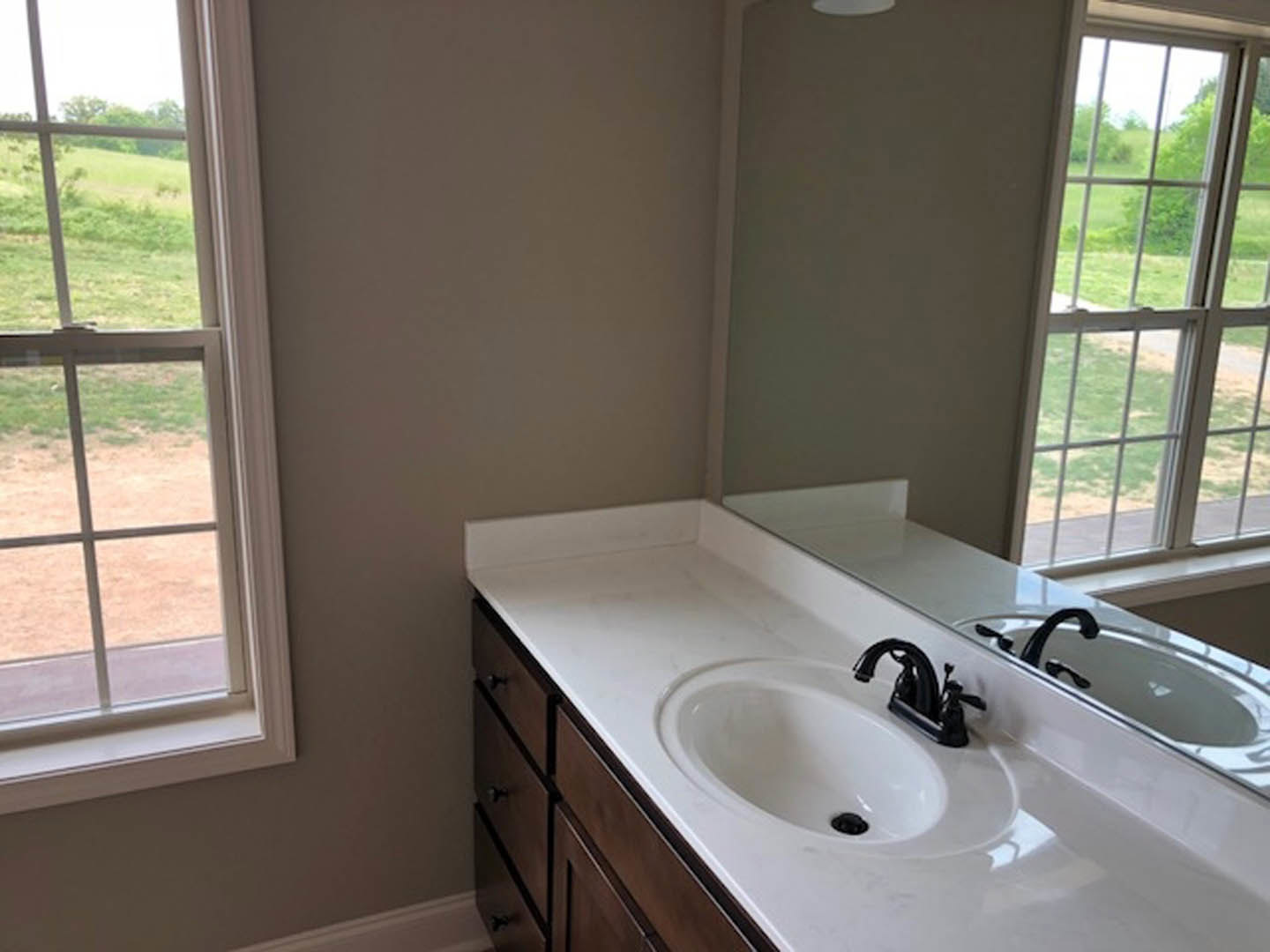 White porcelain sink with chrome faucet, rectangular mirror above, freestanding bathtub nearby, large window overlooking green lawn and trees, light-colored tile walls
