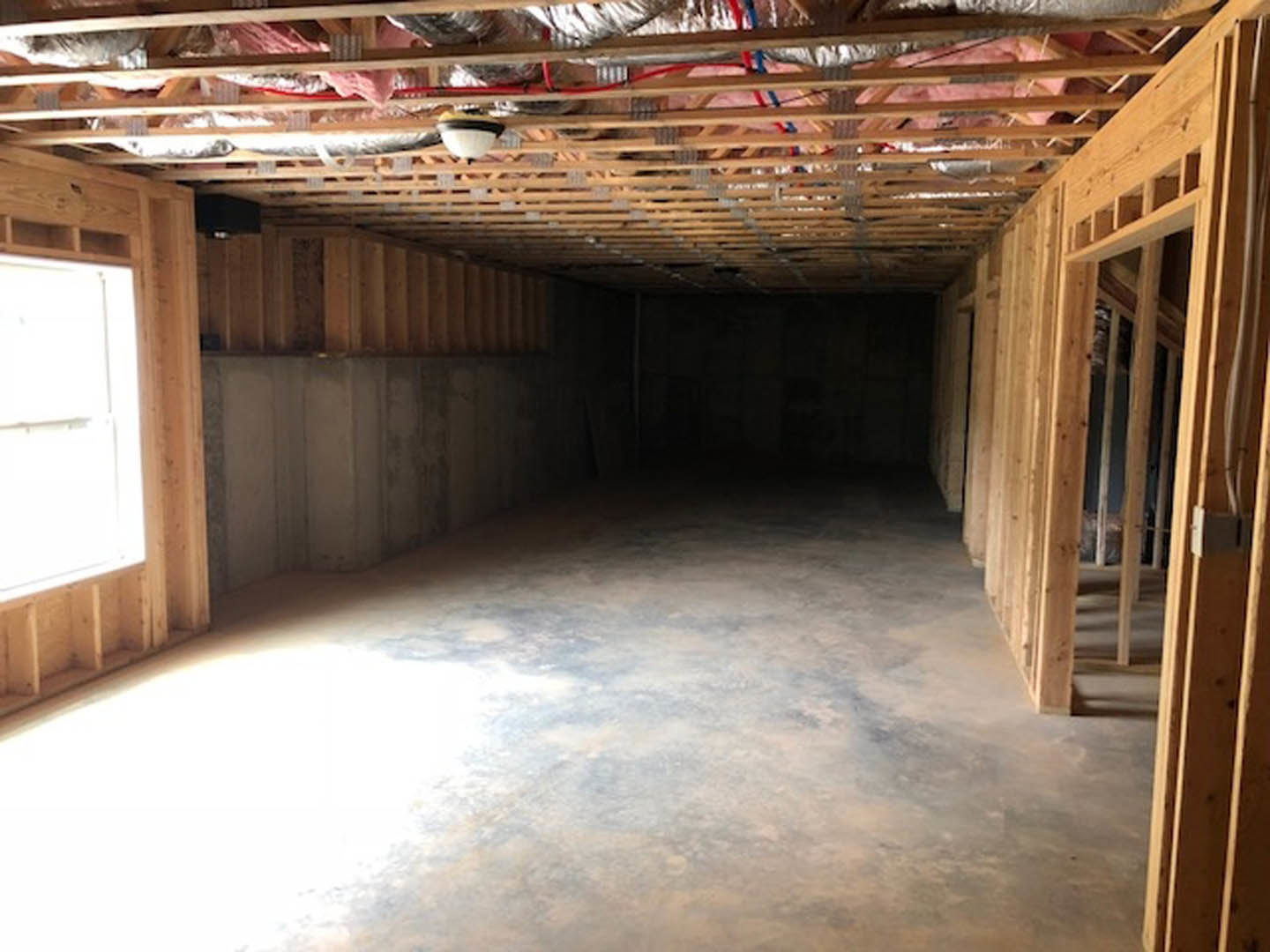 Open room with exposed wood ceiling beams, concrete wall, white door with brown frame, concrete floor illuminated by overhead light, wood framing with visible metal rods and wires