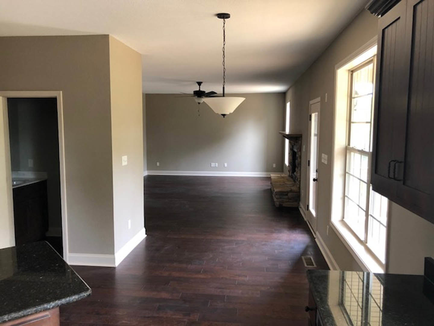 Living room with dark wood flooring, white plaster walls, ceiling fan, black countertop, and a window; door and metal fence details visible