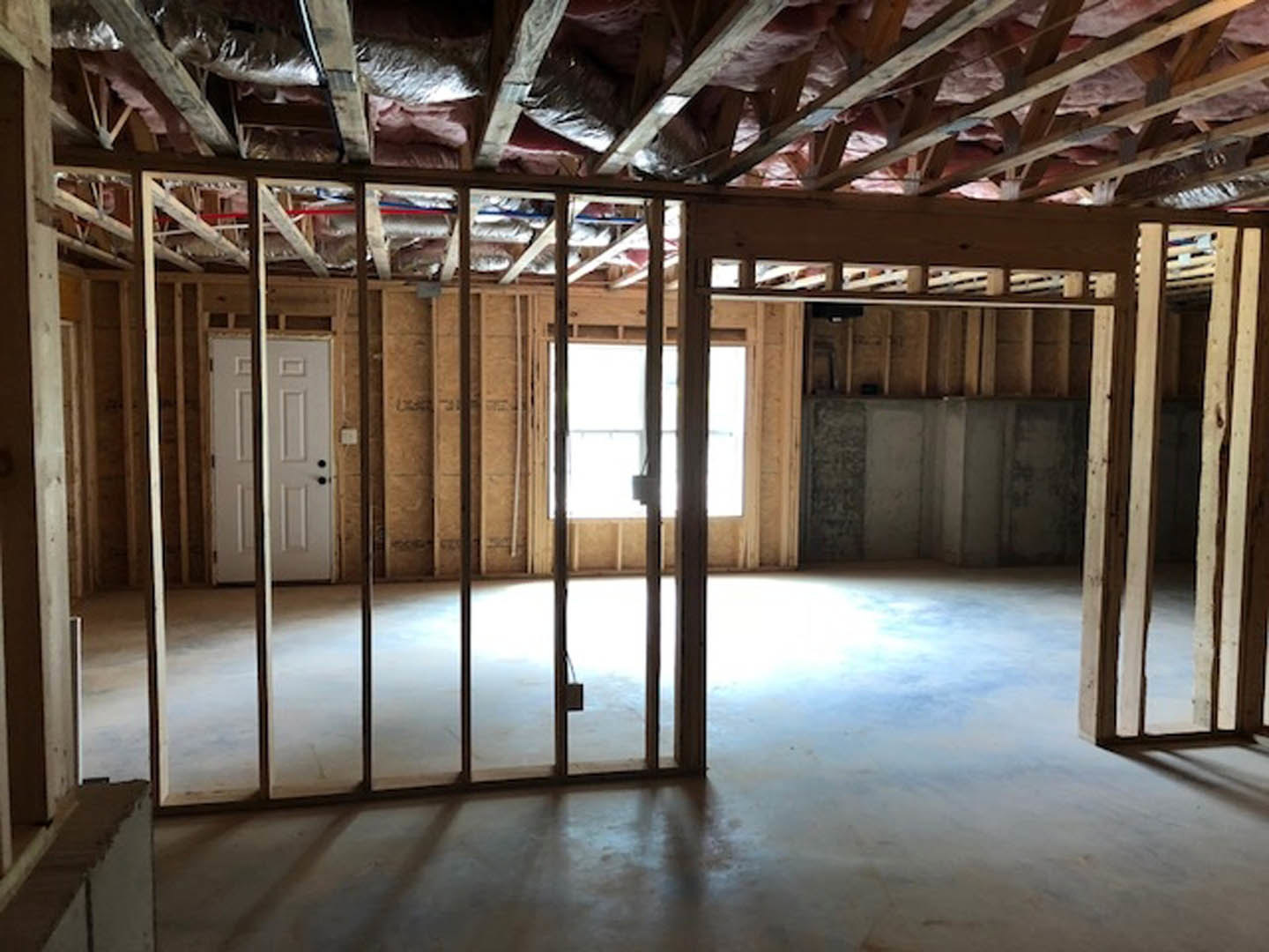 White paneled door with black hardware, adjacent to a window framed in white trim, light streaming onto light wood flooring, exposed ceiling beam and ductwork visible.