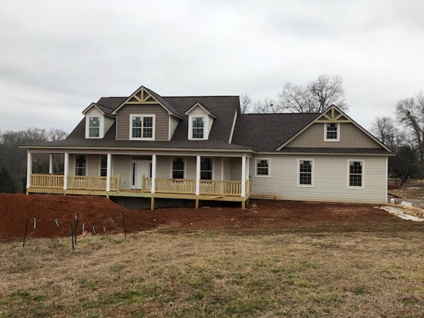 Spacious front porch with wooden decking, white entry door, grid-style windows, manicured lawn bordered by a fence, mature trees, and partly cloudy sky
