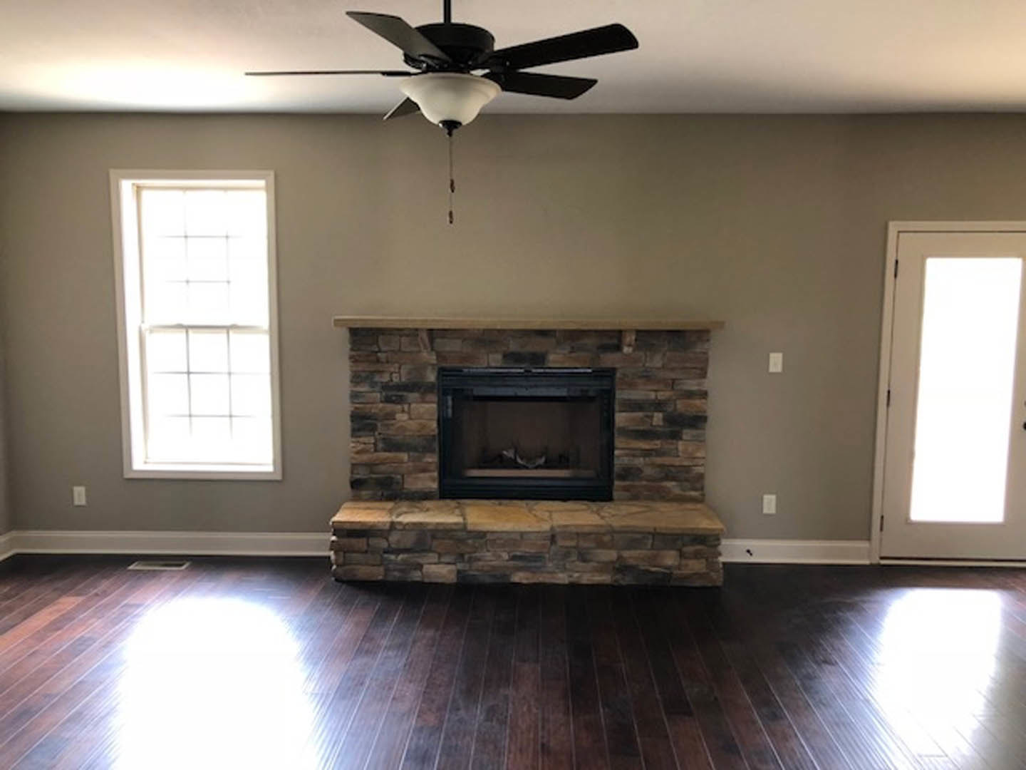 Living room with dark wood floors, stone fireplace, ceiling fan with light, large window, and neutral walls