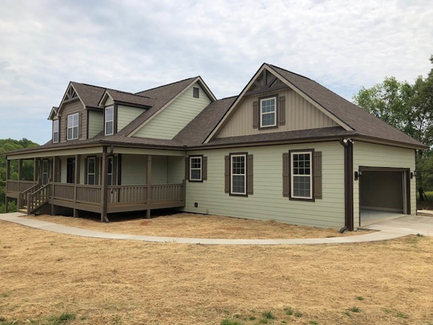 Two-story home with gray siding, white trim, covered front porch, concrete driveway, and large windows; landscaped yard with trees under partly cloudy sky