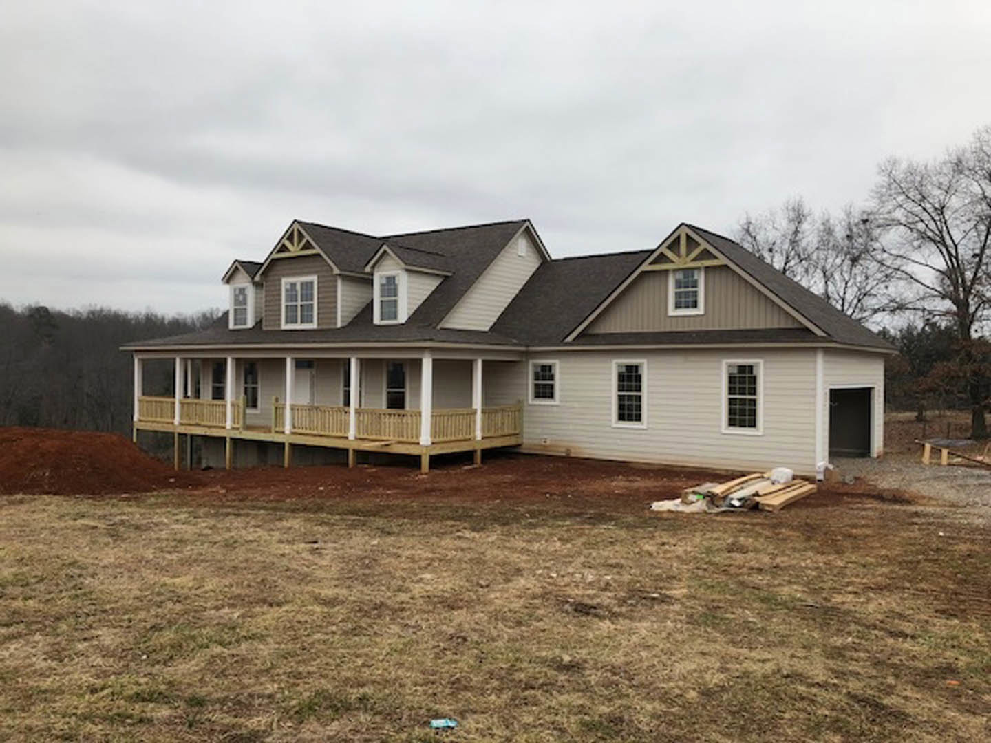 Partially built house with exposed wooden framing, multi-pane windows, covered porch supported by pillars, and expansive grassy yard under a cloudy sky