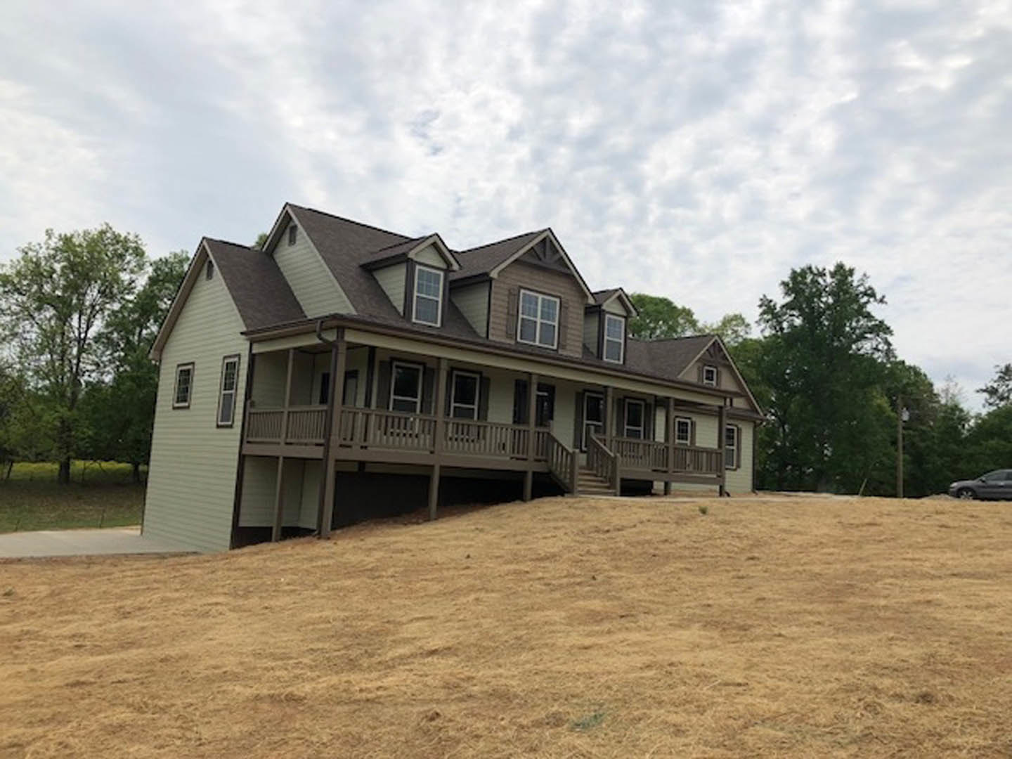 Two-story house with white-framed windows and covered porch, situated on a grassy hill with stone steps, leafy tree in foreground, cloudy sky above.