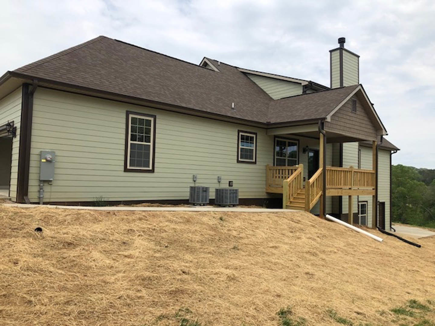 Two-story house with white-framed windows, gray siding, wooden porch and deck, wooden railing, grassy hill, sidewalk, and two trash cans in front.
