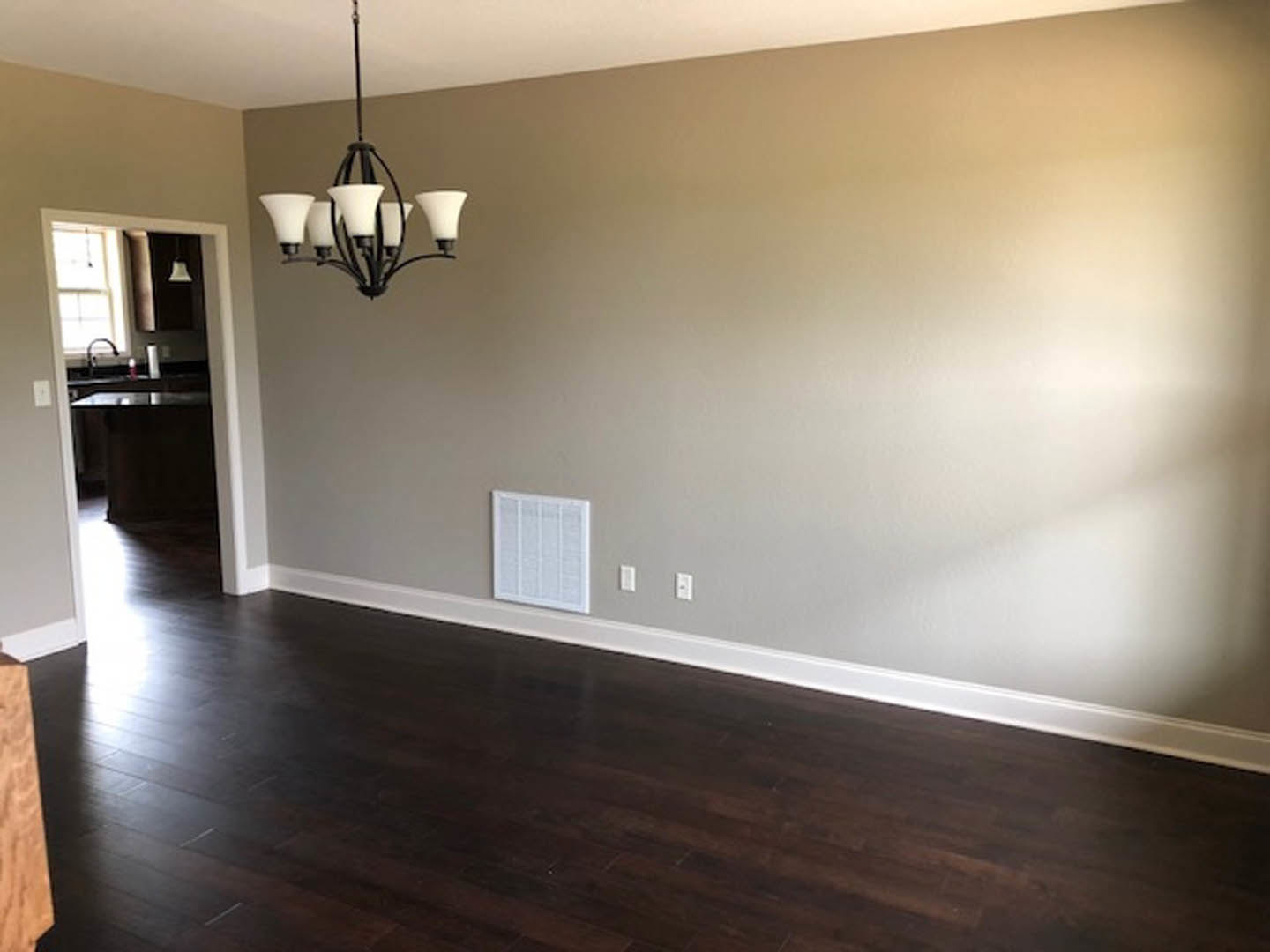 Hardwood floor with wall vent, white plaster walls, ceiling-mounted chandelier with white shades, partial view of lamp, blurred arm in foreground