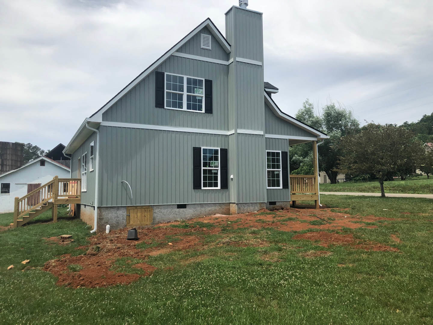 Two-story cottage with multiple windows, wood deck, green lawn, wooden stairs, and fenced yard surrounded by trees and plants under a cloudy sky