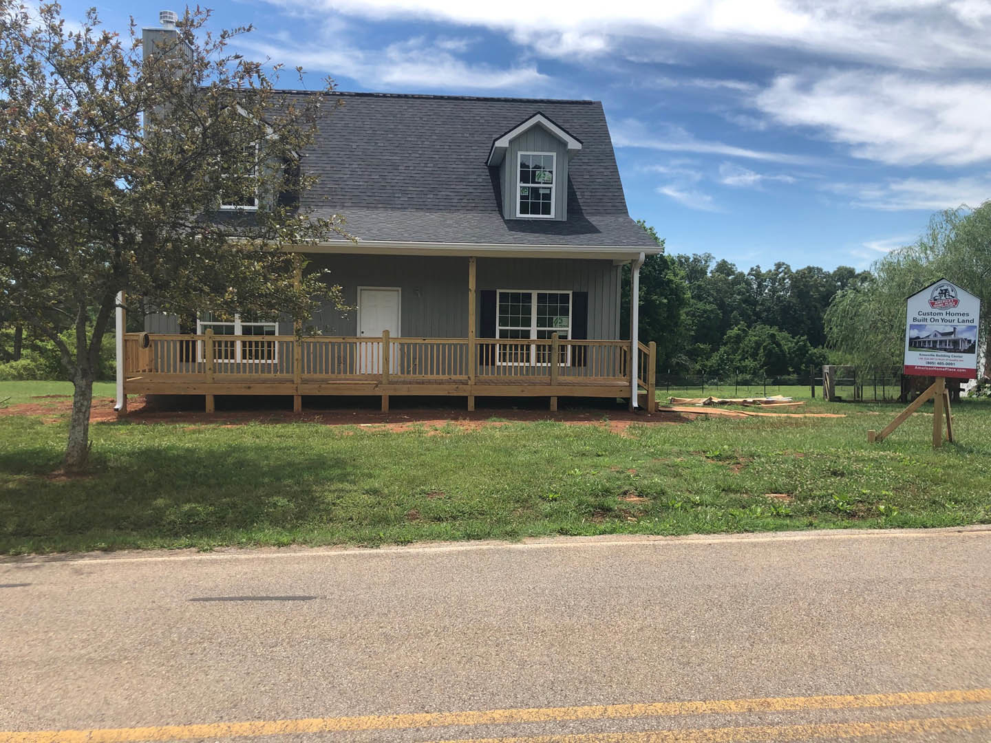 Two-story cottage-style home with white siding, covered front porch featuring wooden railings, multi-pane windows, manicured lawn, mature tree in backyard, and partly cloudy sky