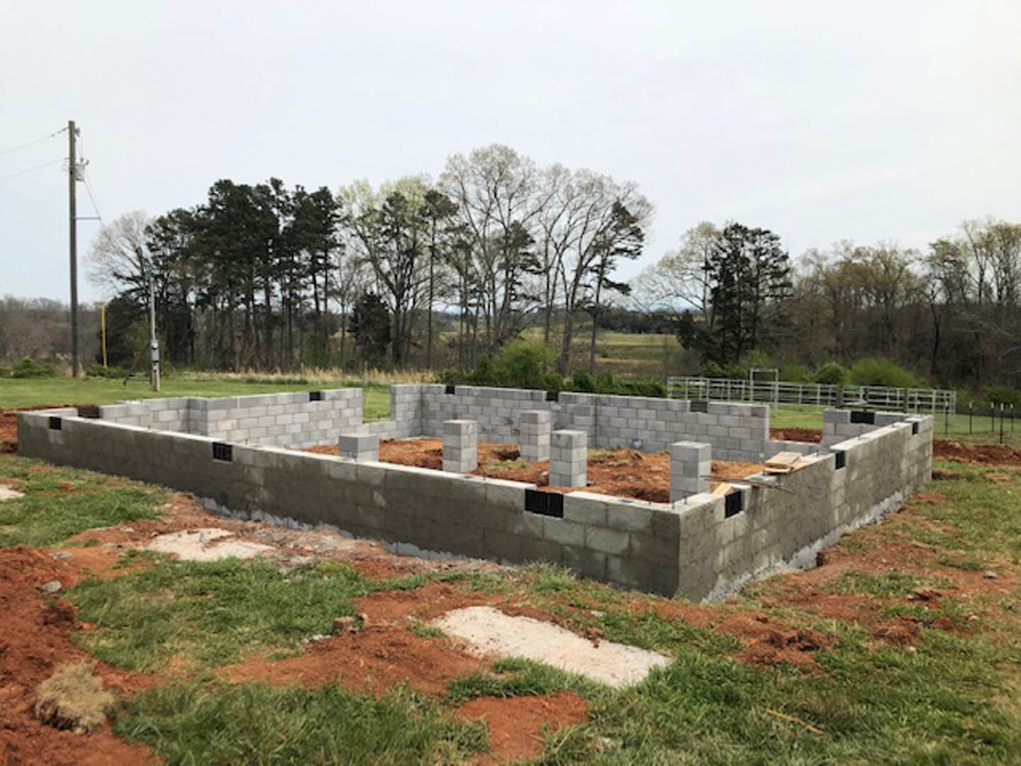 Concrete foundation and excavation surrounded by grassy field, white stone path leading toward construction site, leafless trees in background, street light visible at edge of