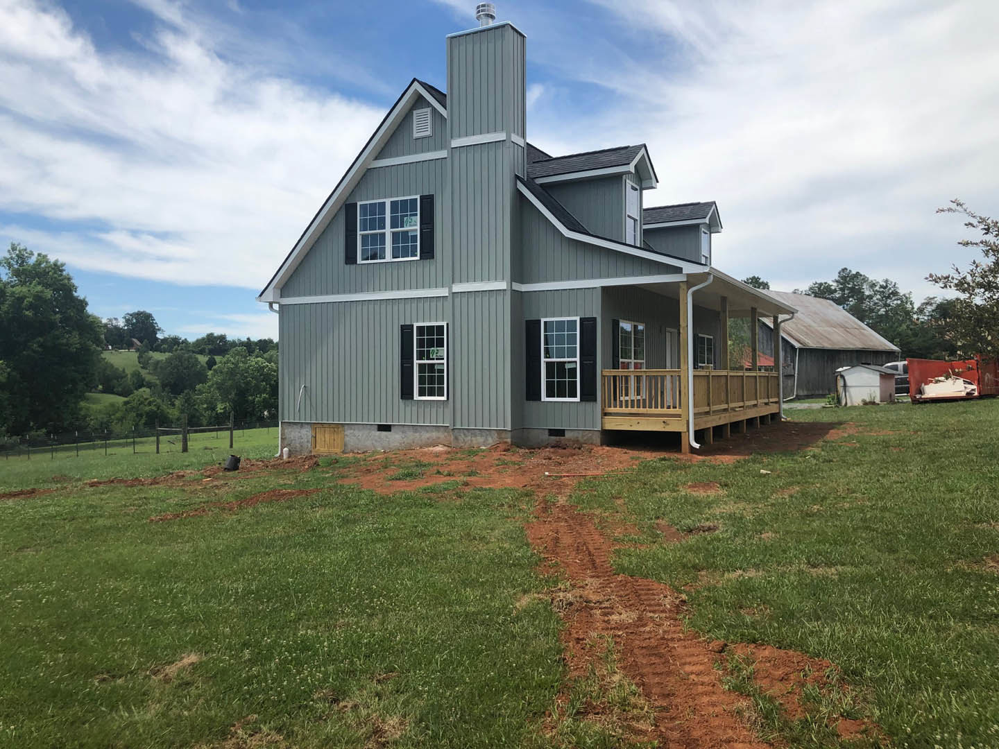 Two-story home with white siding, covered front porch, wooden deck, fenced yard, chimney, grassy lawn, and tire tracks on dirt driveway