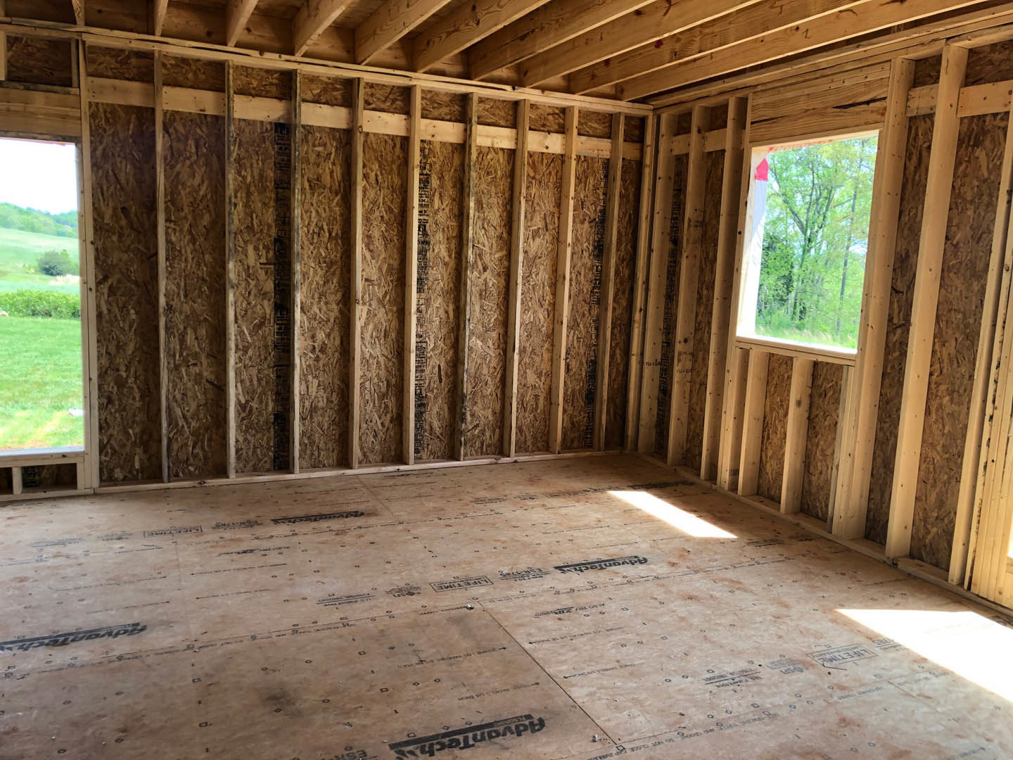 Sunlit room with unfinished wood flooring, exposed ceiling beams, and a large window overlooking green grass and trees.