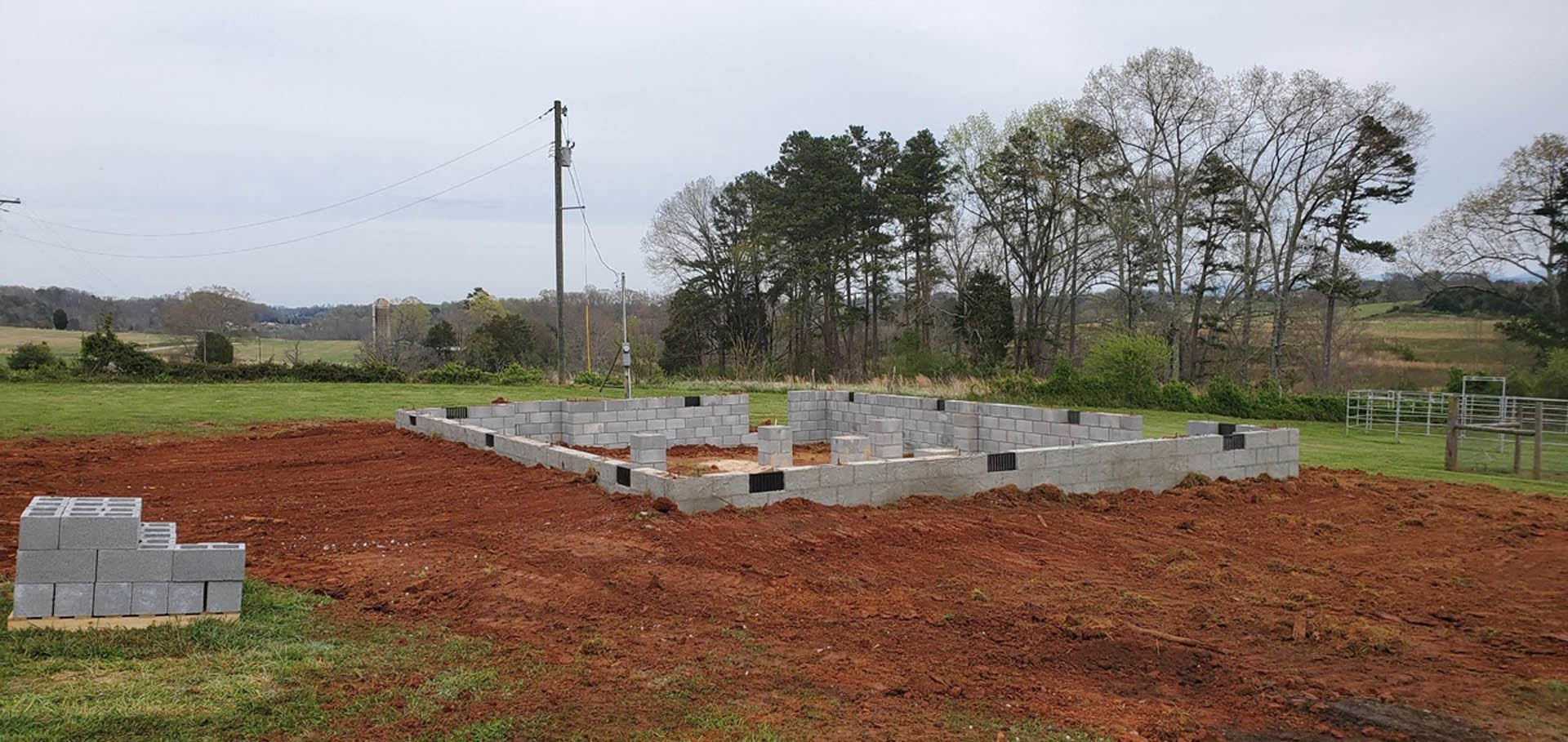 Concrete foundation with stacked grey blocks, fenced construction site, grassy lot, scattered building materials, cloudy sky overhead