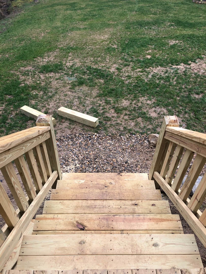 Wooden staircase with plank railing ascending to grassy yard, bordered by wooden fence and scattered lumber on ground