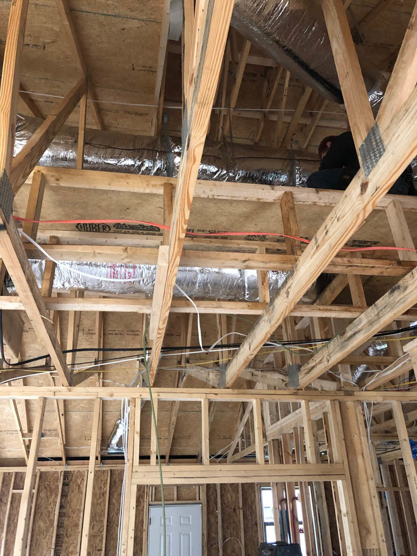 Exposed wood framing and ceiling beams in a partially constructed home interior, with building insulation visible between planks, a white door, potted plant in the background