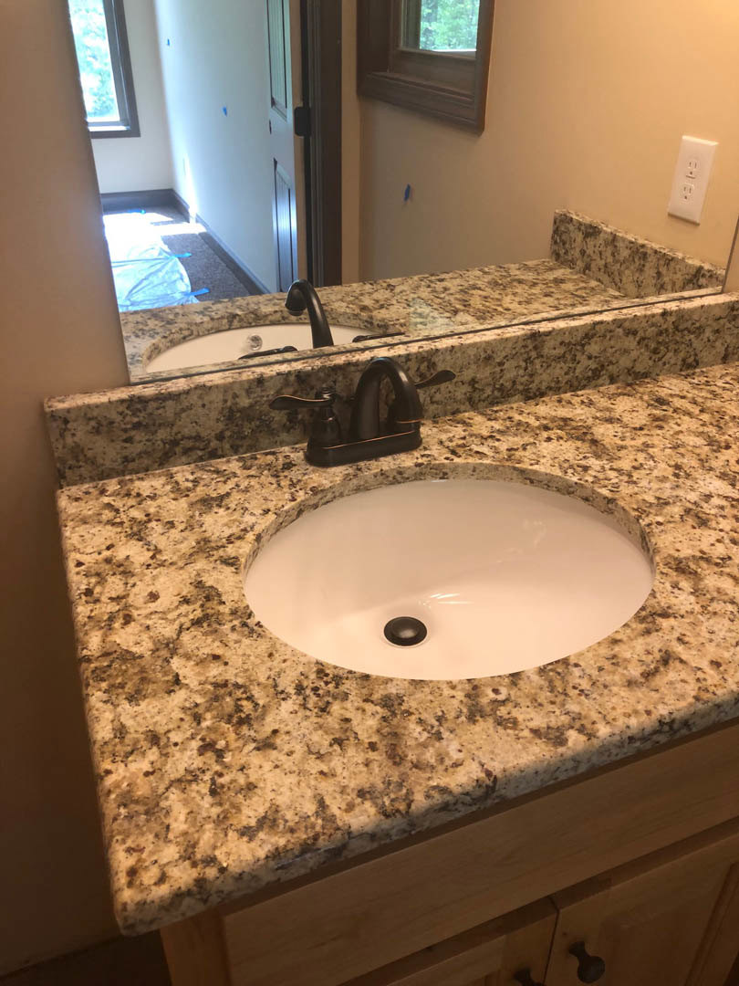 White rectangular sink with black drain and chrome faucet set in a light stone countertop, large frameless mirror above, white tile backsplash, electrical outlet, and window with