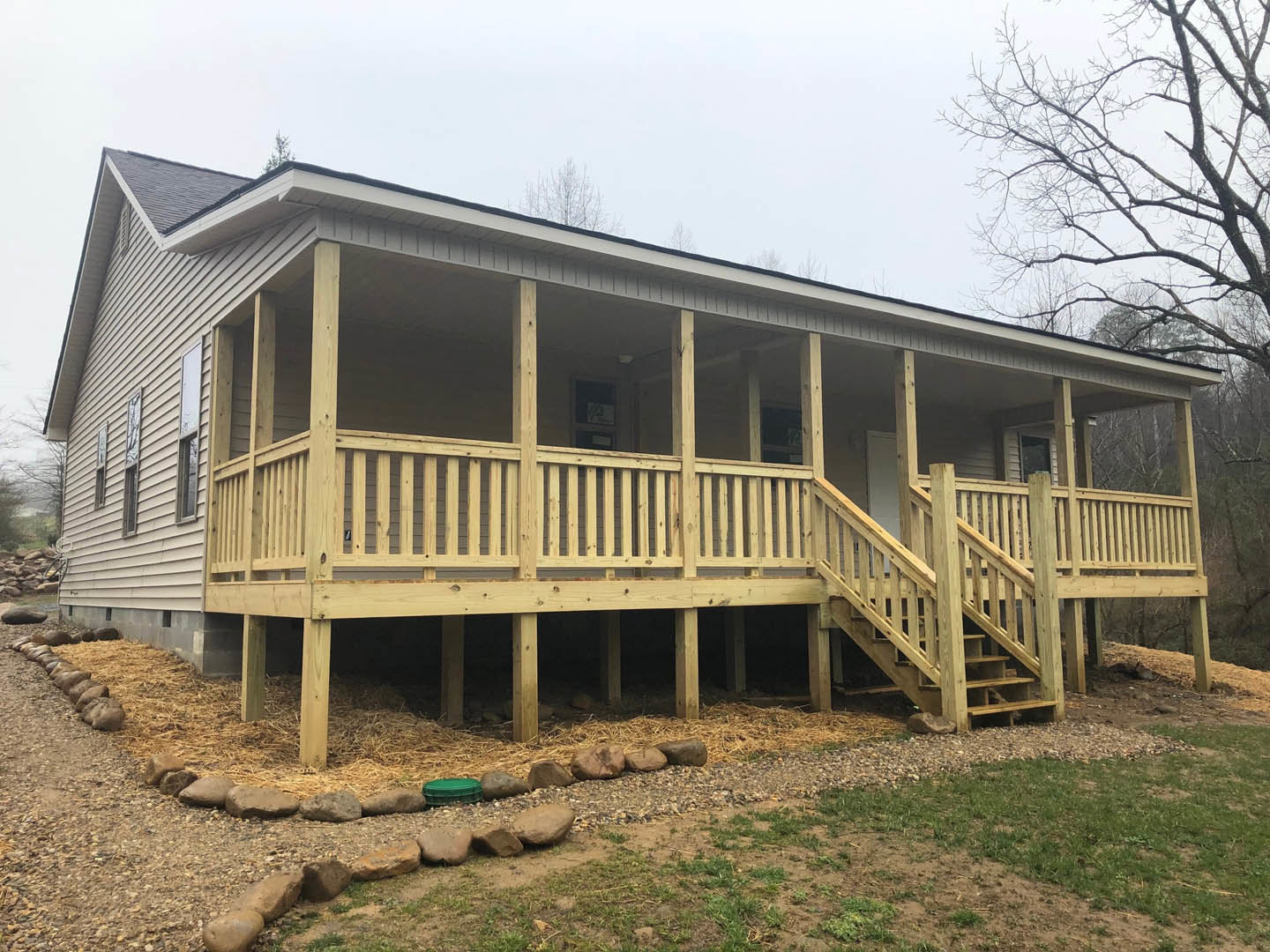 White siding house with covered front porch, wooden railing, and picket fence; leafless tree and rocks line dirt path in landscaped yard.