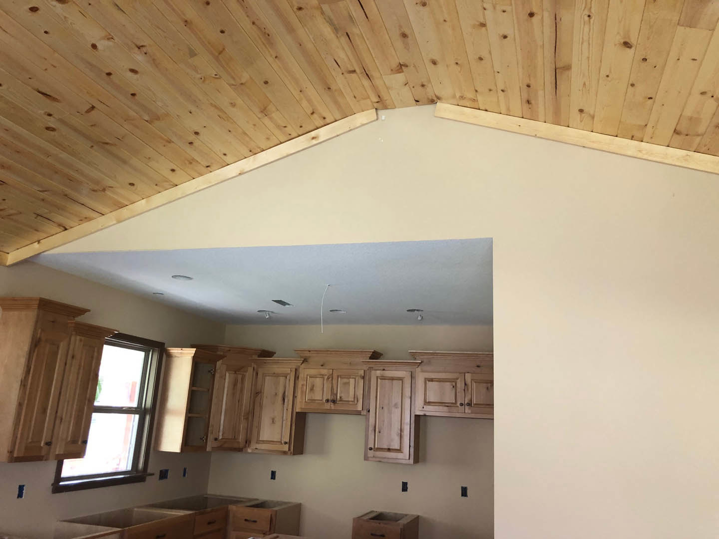 Kitchen with wood plank ceiling, wood paneled wall, window featuring wooden shutter, white plaster walls, close-up of a door, brown storage box with black handle, exposed ceiling