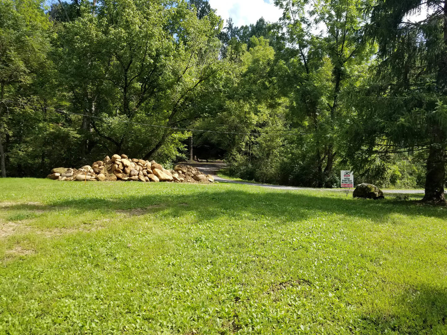 Grassy lawn with a pile of rocks surrounded by mature trees under a clear sky