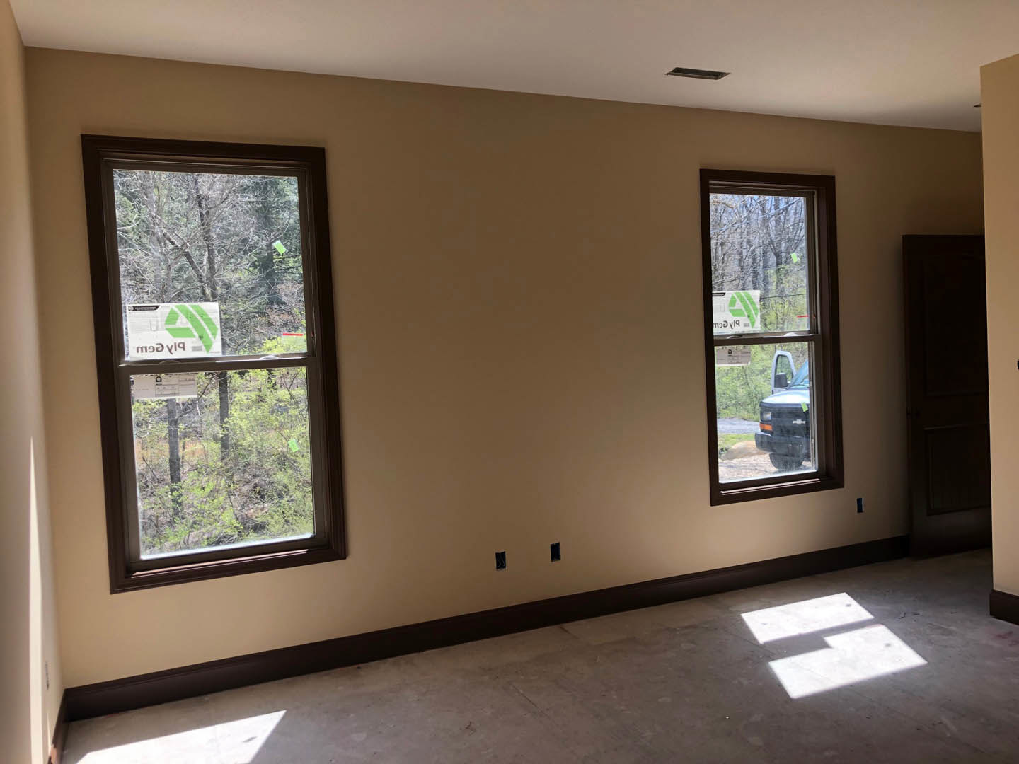 Unfinished room with concrete floor, two windows, exposed electrical outlets on white wall, construction sign visible through window, parked truck outside, portable work light on