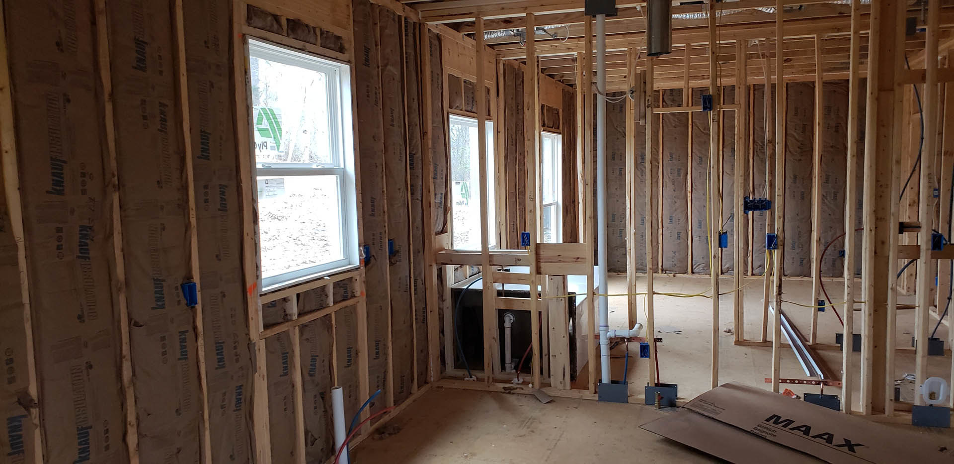 Unfinished room with exposed wood framing, multiple windows, visible pipes, insulation, and construction materials including a box and a white container with gold coins.