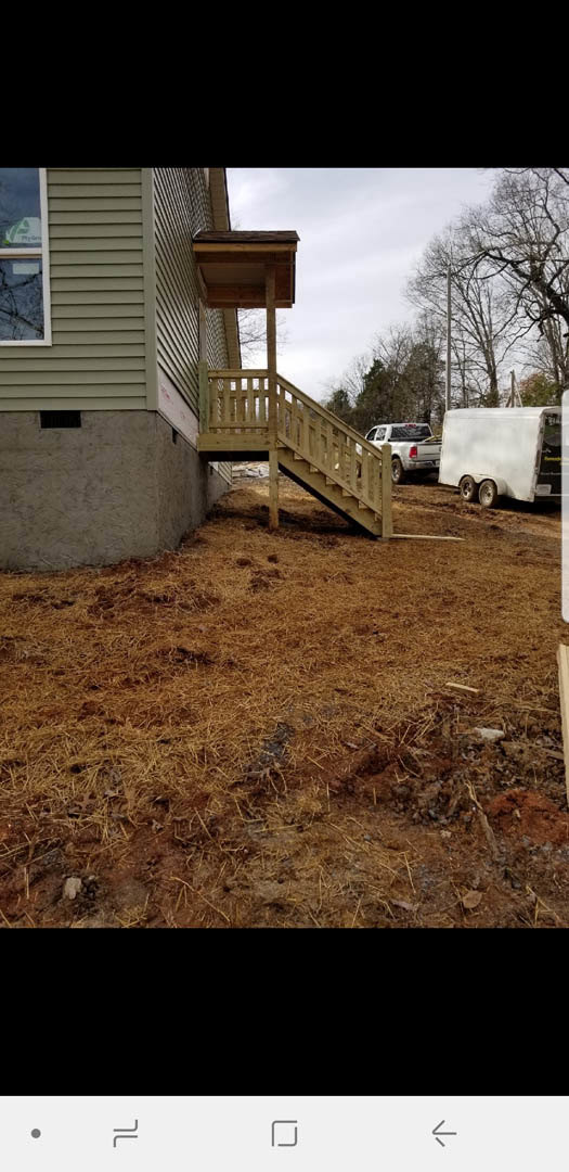 Wooden staircase leading to entry of custom home, white truck parked on dirt driveway in back, straw-covered ground, windows visible on exterior, trees in background.