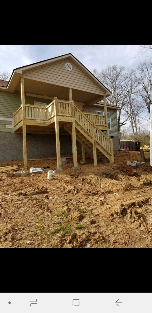 Wooden deck with railing and staircase leading to light-colored siding home, surrounded by mature trees under partly cloudy sky