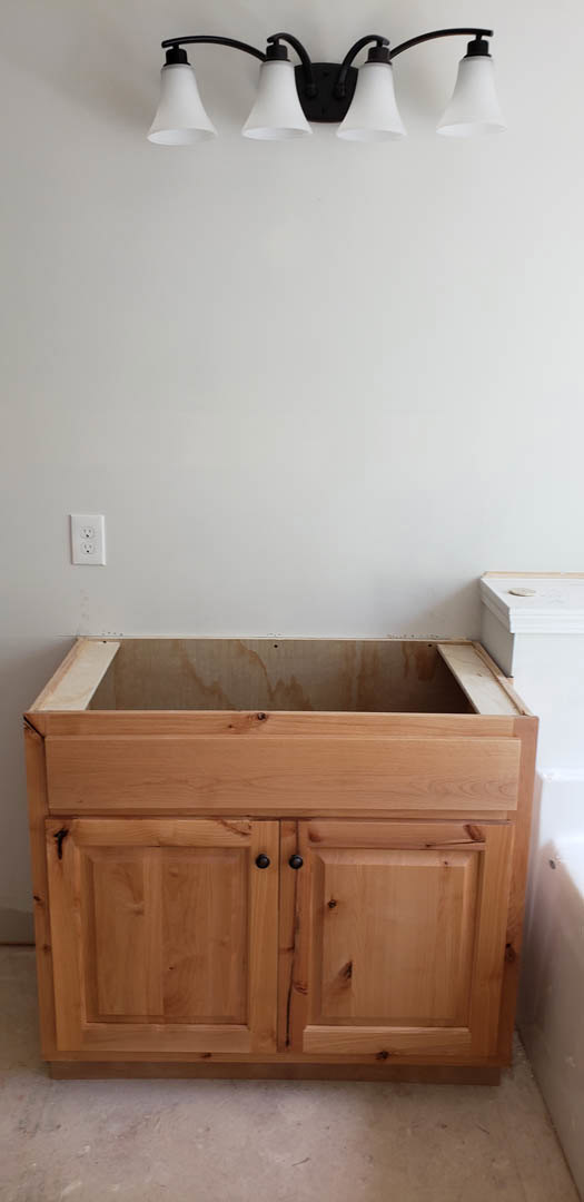 Light wood bathroom cabinet with a cutout in the top, two white lamp fixtures above, brown stain visible on the wall, white sink and faucet nearby, tiled floor.