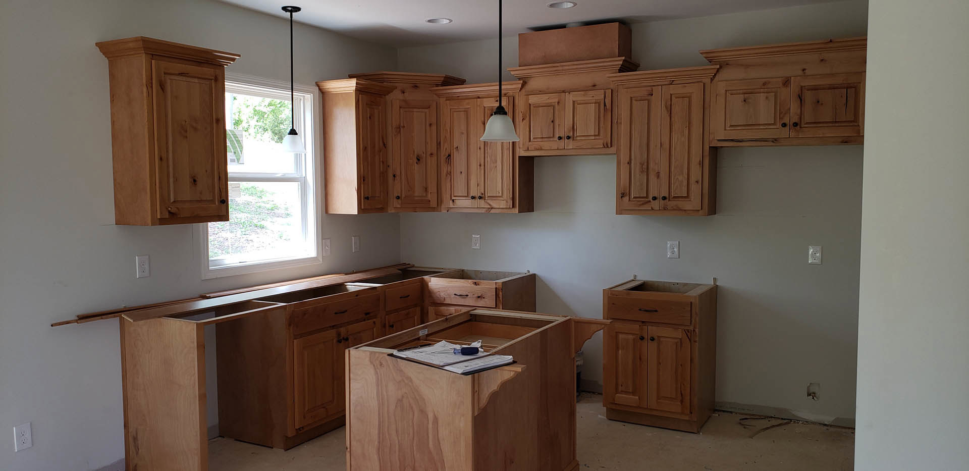 Kitchen with natural wood cabinets, stone countertop, built-in drawers, window above sink, white electrical outlet on wall, black metal support pole near cabinetry, clipboard and