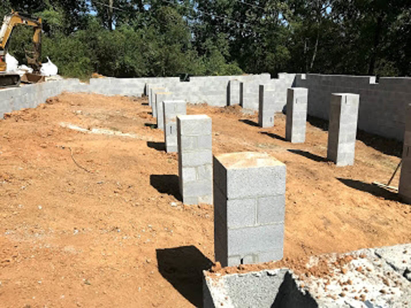 Stacked concrete blocks on a dirt construction site, white brick wall with black metal frame, scattered rocks and soil, tree trunk visible in background, shadow cast across ground