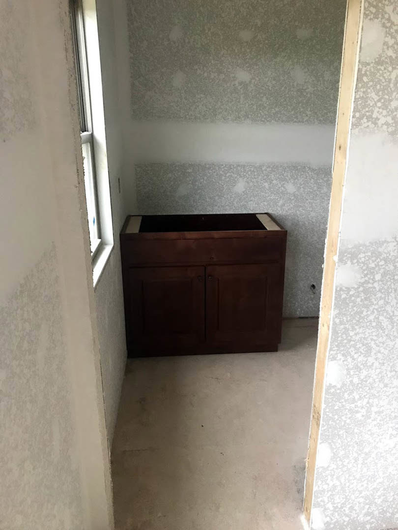 Brown wood cabinet with sink beneath a window, white floor, open door, plaster walls, black rectangular object with white border, broken windowpane
