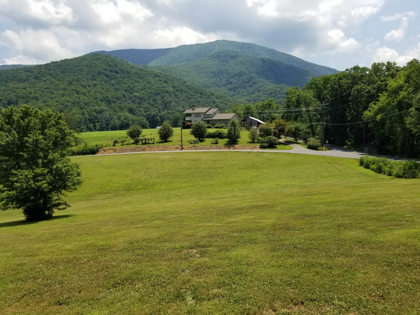 Expansive grassy field bordered by leafy trees, circular walkway, and a custom home set against a mountain backdrop under a partly cloudy sky