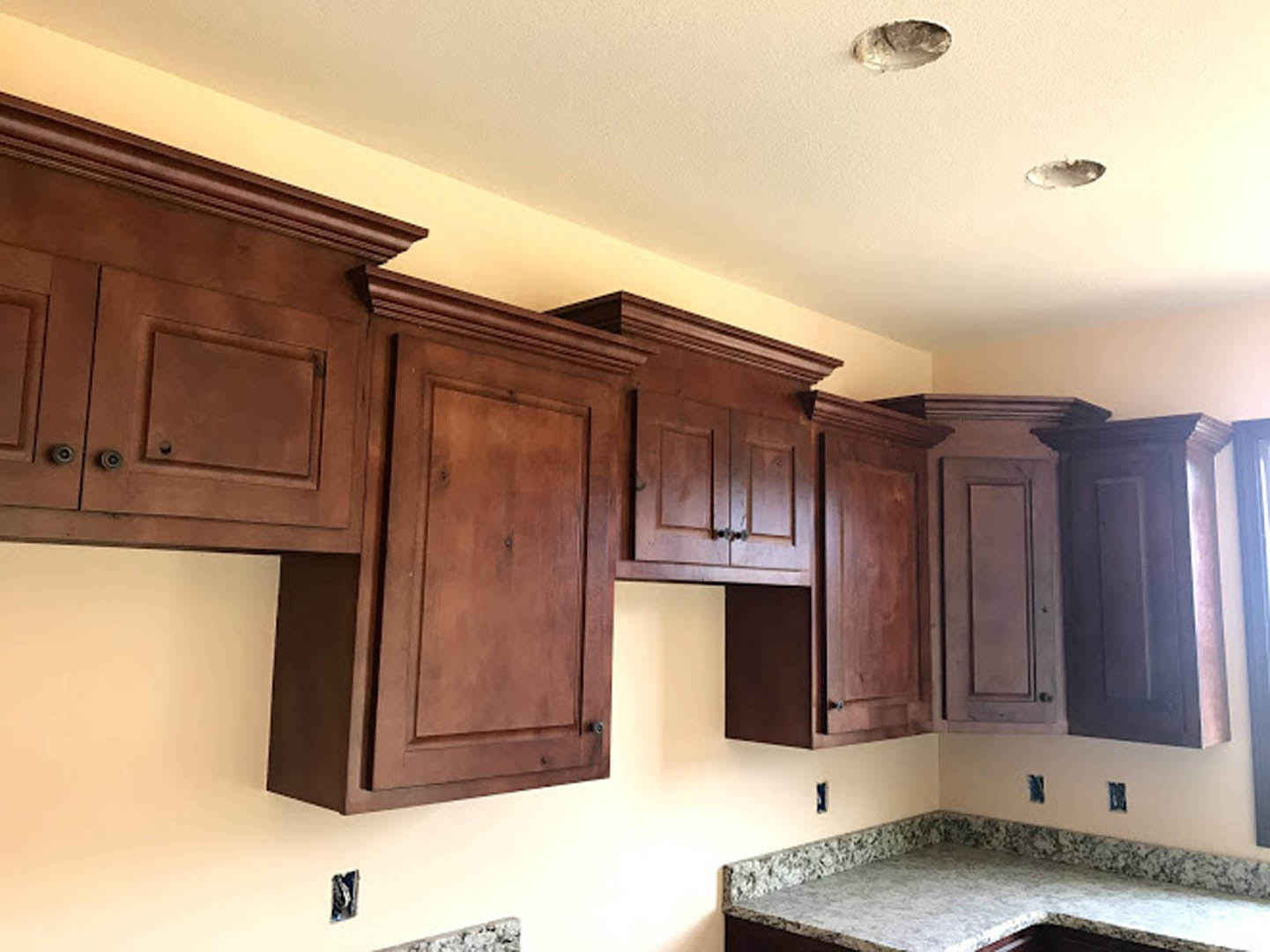 Kitchen with light wood cabinets, marble countertop, upper cabinets above counter, stainless steel sink, window with trim, and wooden door featuring circular cutout