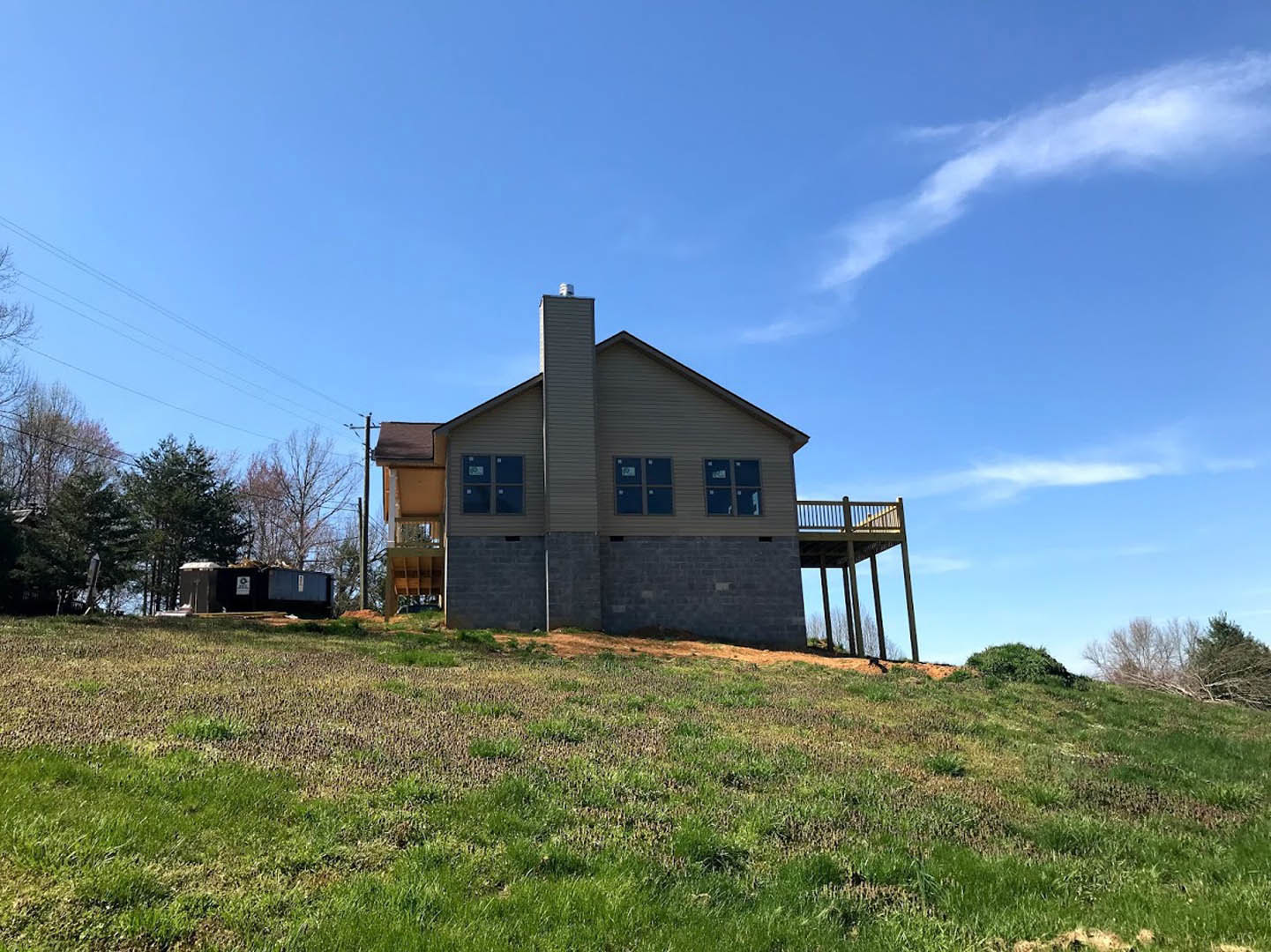 Two-story house with gray siding and white trim, elevated on a grassy hill, featuring a wooden deck and covered porch, brick retaining wall along the lawn, blue sky with scattered