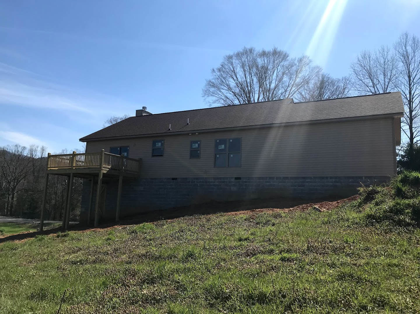Two-story house with brick exterior, large windows, and elevated wooden deck overlooking grassy hillside, surrounded by trees and open sky