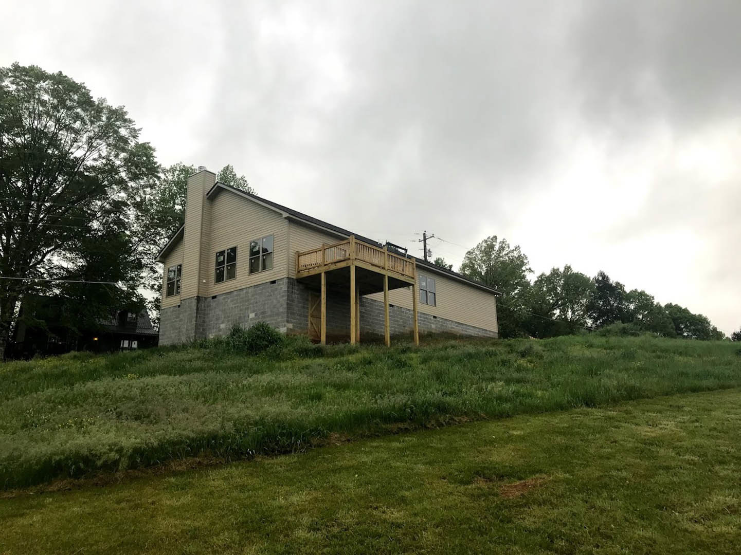 White farmhouse with covered wooden porch on grassy hill, mature leafy tree beside house, large windows, cloudy sky overhead