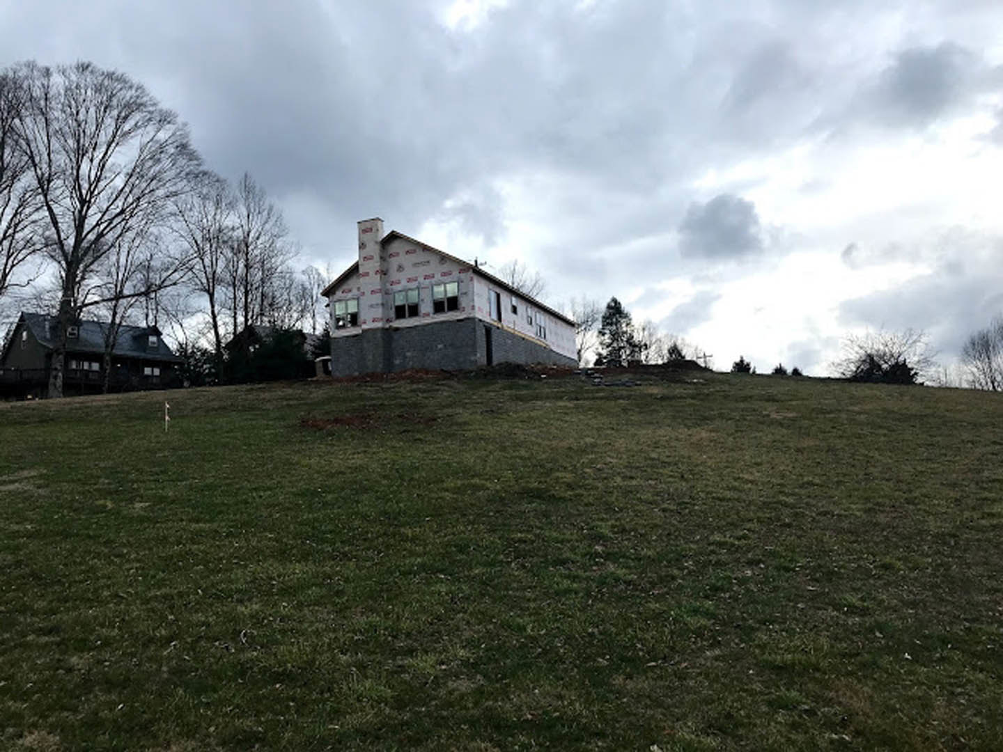 Modern house under construction on grassy hillside, surrounded by trees, with white pillars marked by red tape and cloudy sky overhead