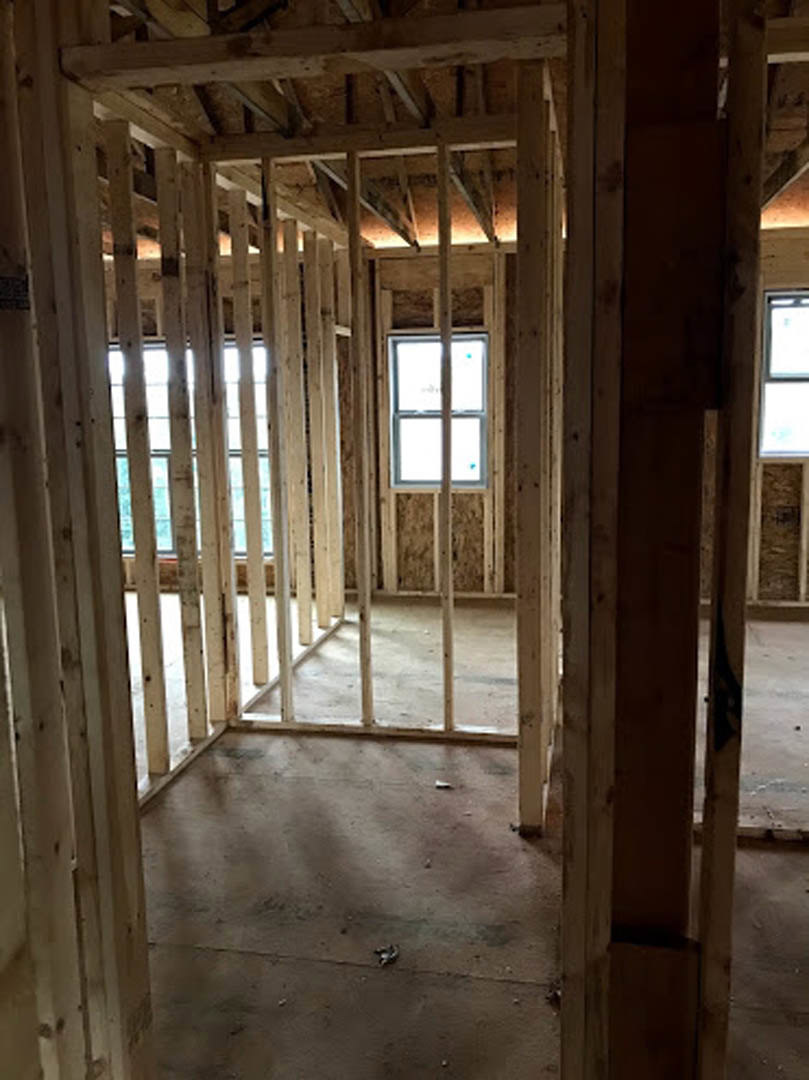 Unfinished room with exposed wood framing, ceiling beams, and a window displaying a sign; daylight streams through the window, highlighting construction materials and insulation.
