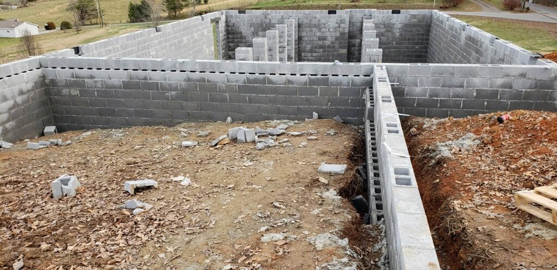 Concrete foundation with exposed hole in soil, wooden pallet on dirt slope, pile of bricks, close-up views of concrete wall, stone, and concrete block, scattered rubble and grass.