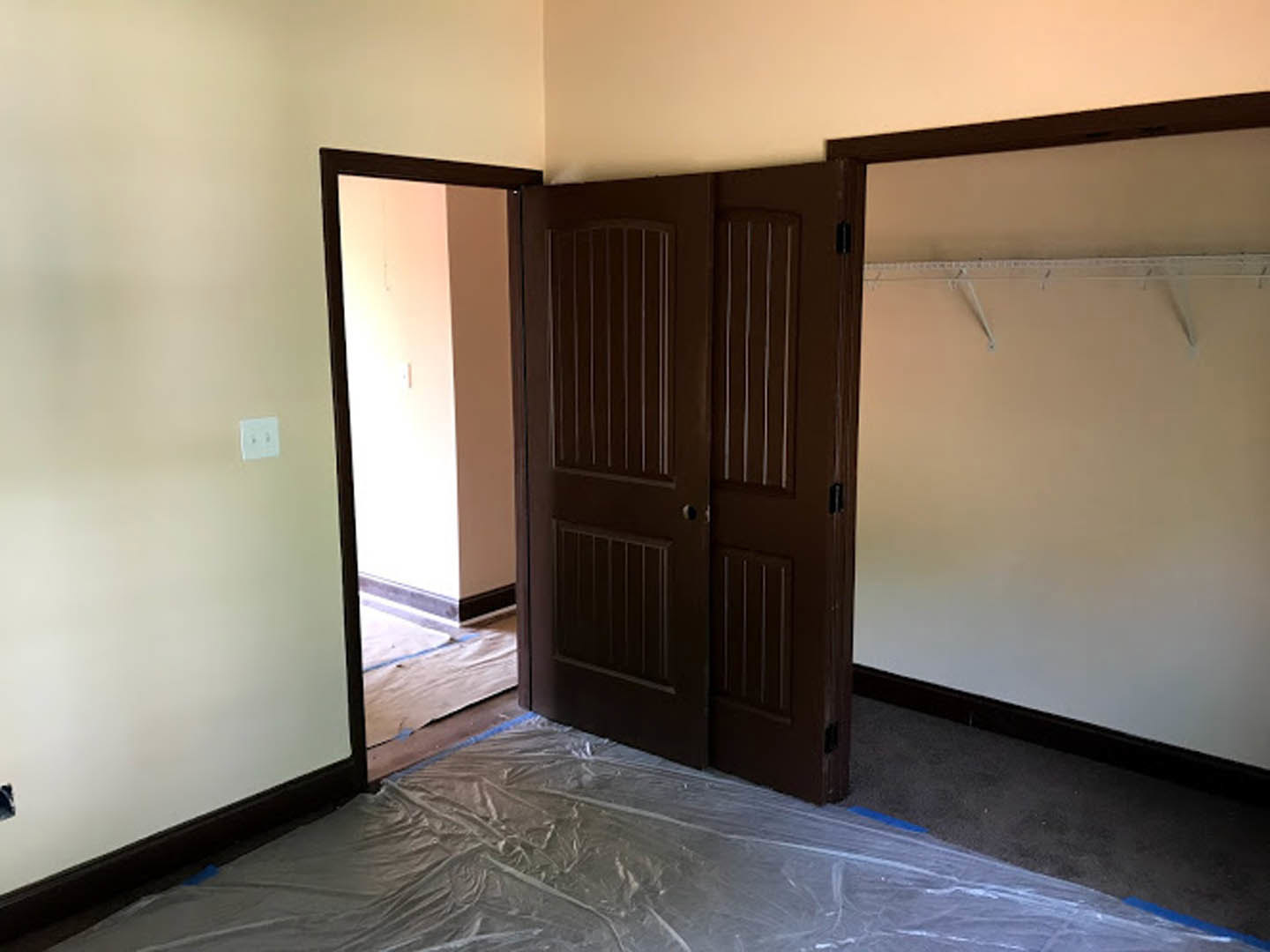White-walled room with two brown doors, black door knobs, built-in white shelves, light switch, and plastic covering the floor