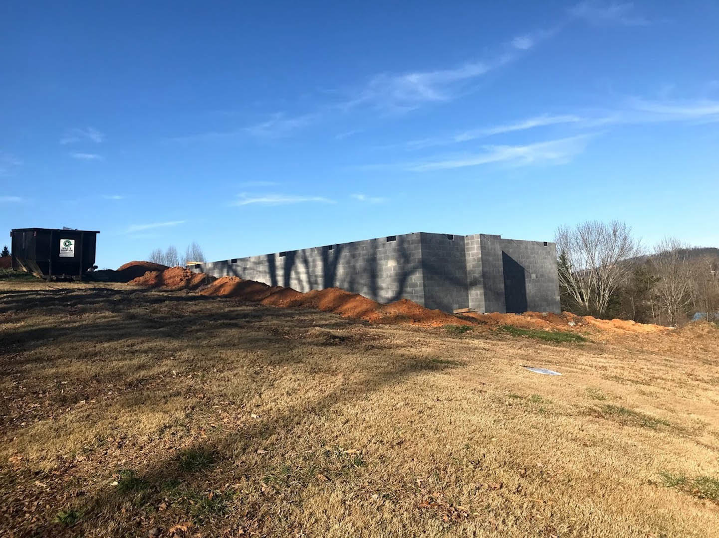Modern home exterior with brick and siding walls, large pile of dirt beside the foundation, leafless tree, grassy field, and blue sky with scattered clouds