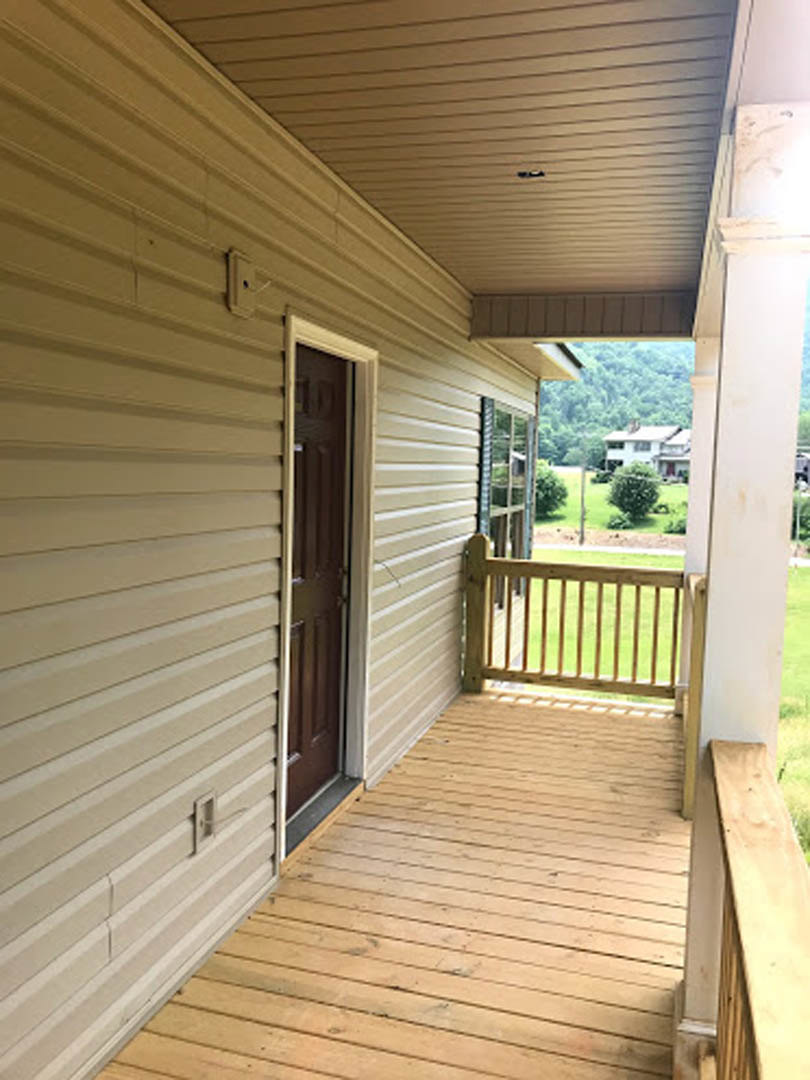 Brown front door on a wooden porch with matching railing and deck, light-colored siding exterior