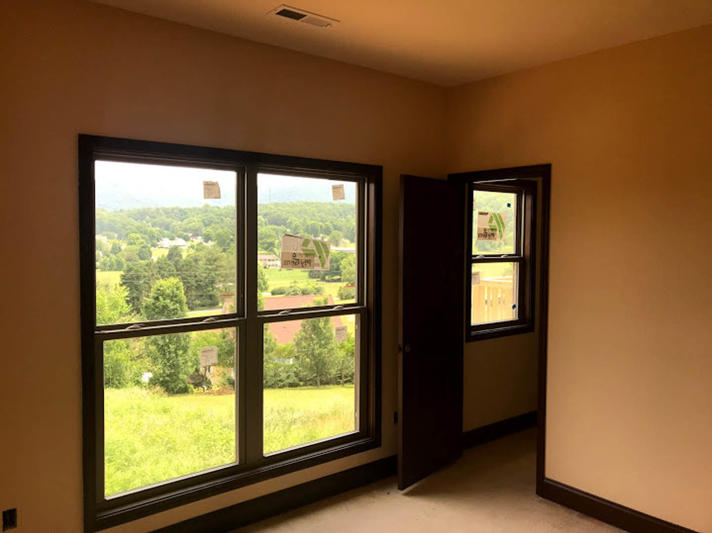 White-walled room with wood floor, single window showing field and trees, closed door, ceiling vent, electrical box, window with taped sign, natural daylight illuminating interior