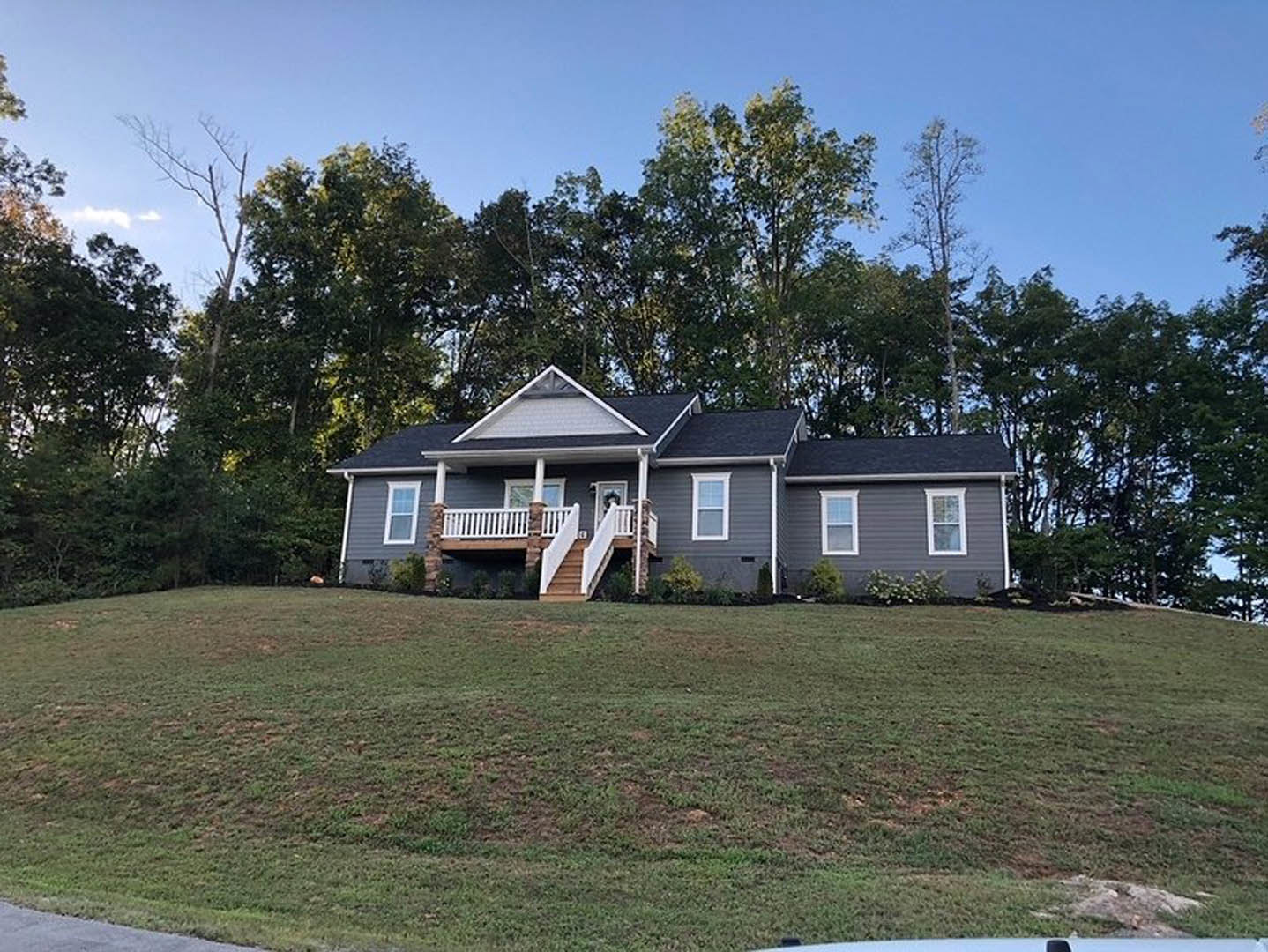 Two-story house with white porch railing, central staircase leading to entry, surrounded by green lawn and mature trees in the background